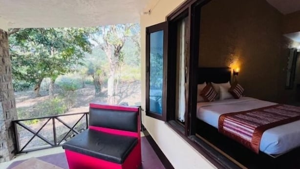 A room with a bed, a window, a balcony with a red chair, and a view of trees. - The Harikanta Corbett Resort (Marchula)