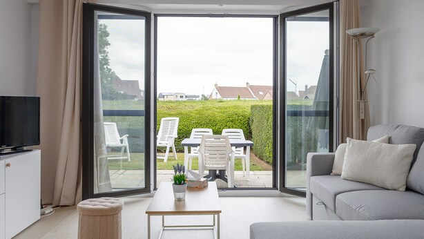 A modern living room with a sofa, a TV, a table with a plant, and a view of a garden through large glass doors. - Ground Floor Apartment With Sunny Terrace (Middelkerke)