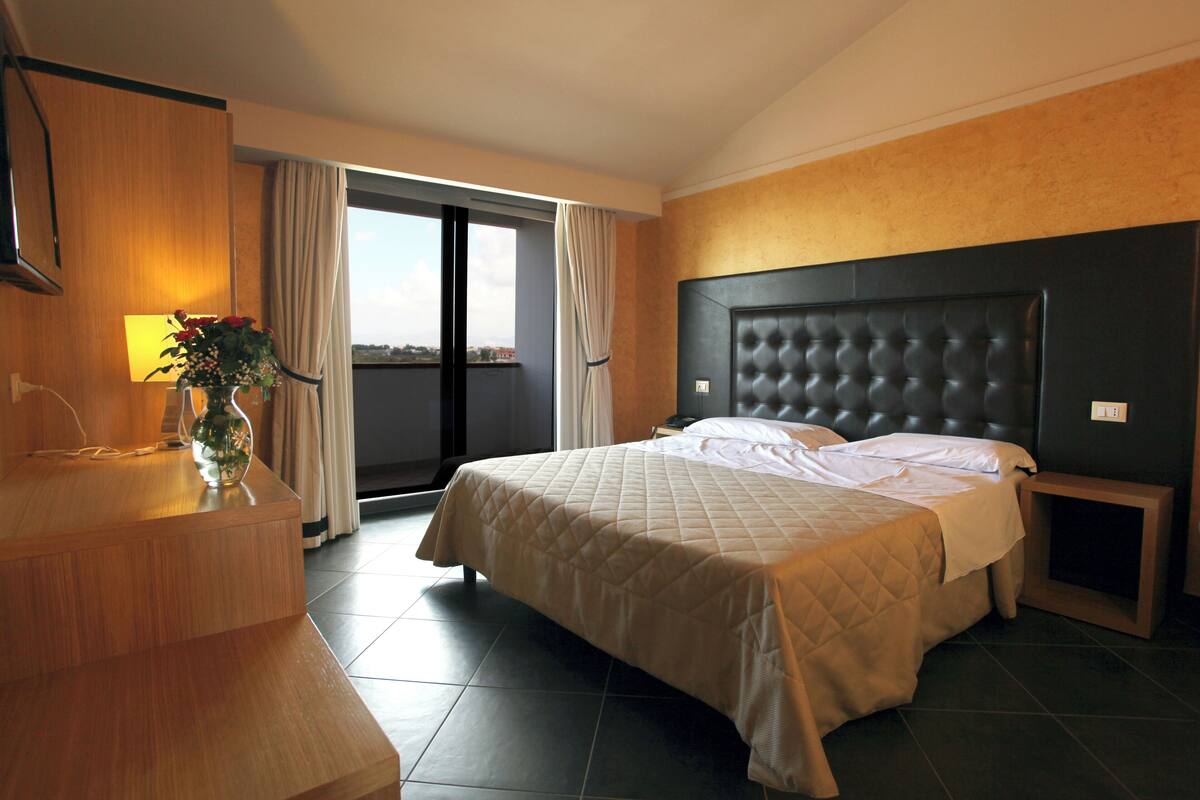 A hotel room with a bed, a bedside table, a lamp, and a view of the outdoors.