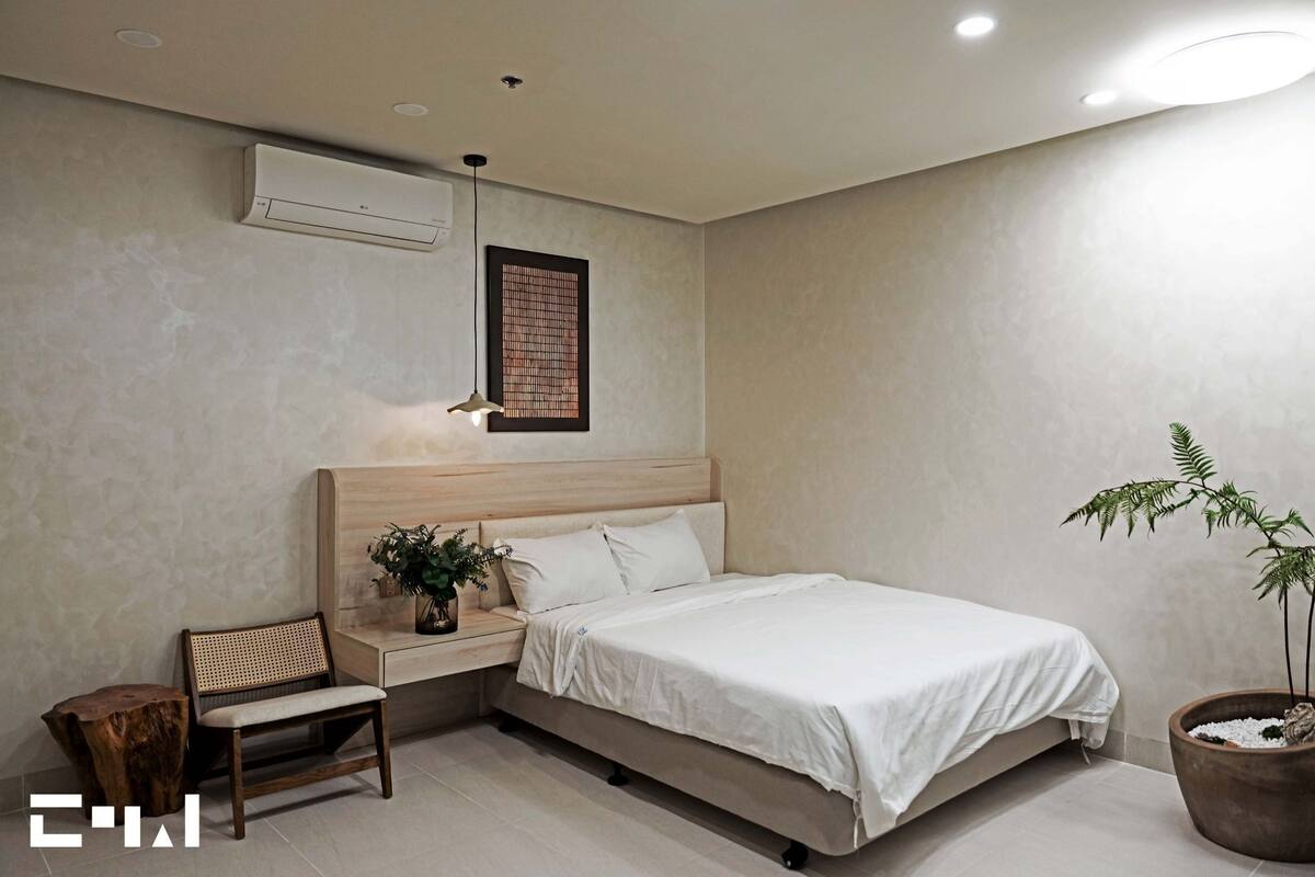 Een slaapkamer met een bed, nachtkastje, stoel, een kamerplant en een wandairconditioner.