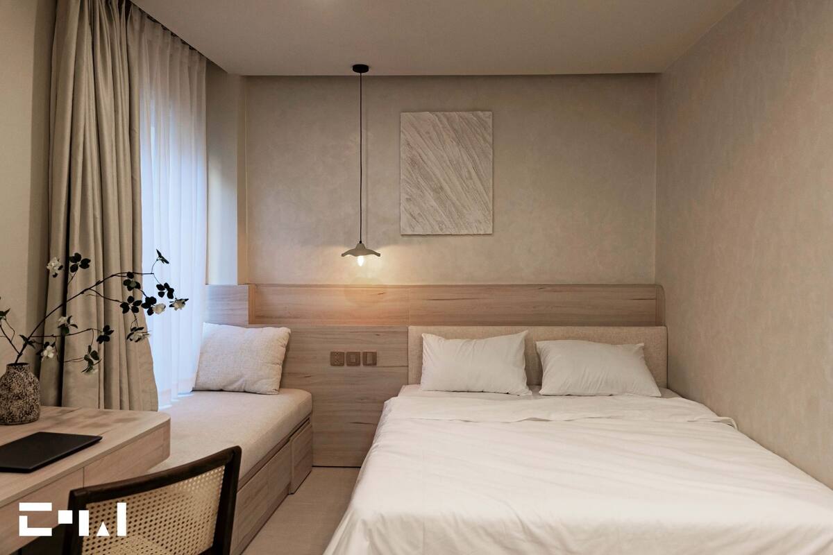 Een moderne hotelkamer met een houten hoofdbord, een nachtkastje met een lamp, een bed met witte lakens en een groot raam met gordijnen.