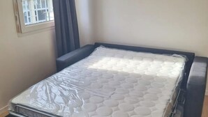 A small, single-bed room with a mattress covered in protective plastic, a window with a view of buildings, and a plain wall.