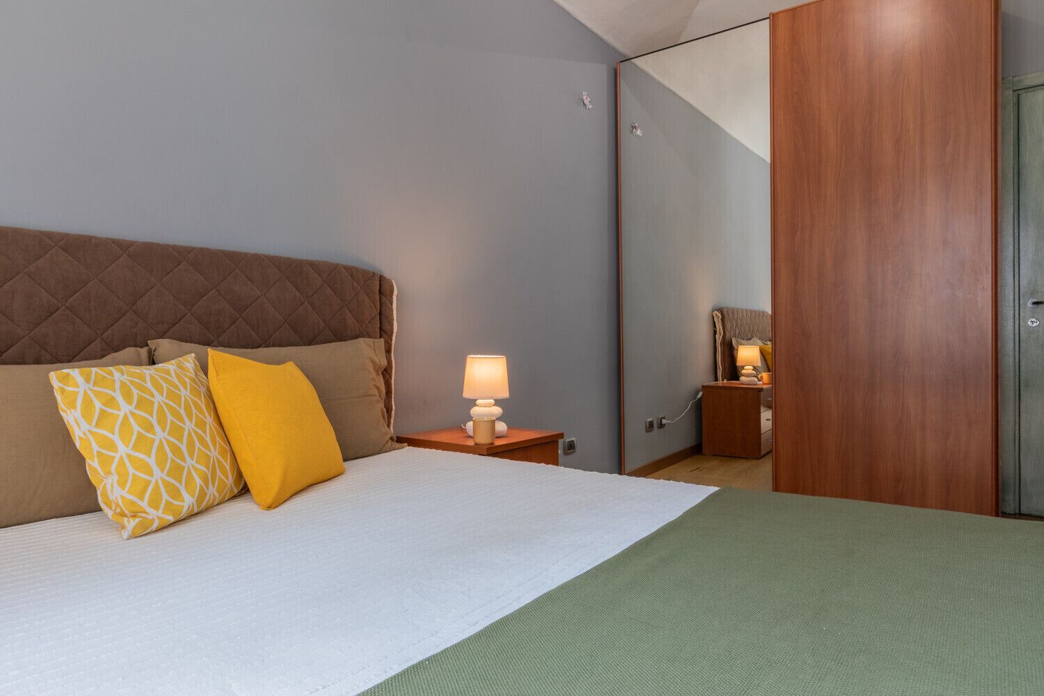 A hotel room with a bed, a nightstand with a lamp, a wooden wardrobe, and a mirror.