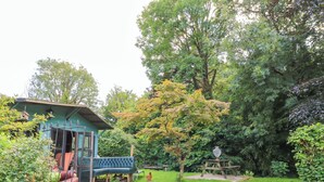 A rustic cabin with a porch and a small deck, surrounded by a lush garden and a large tree.