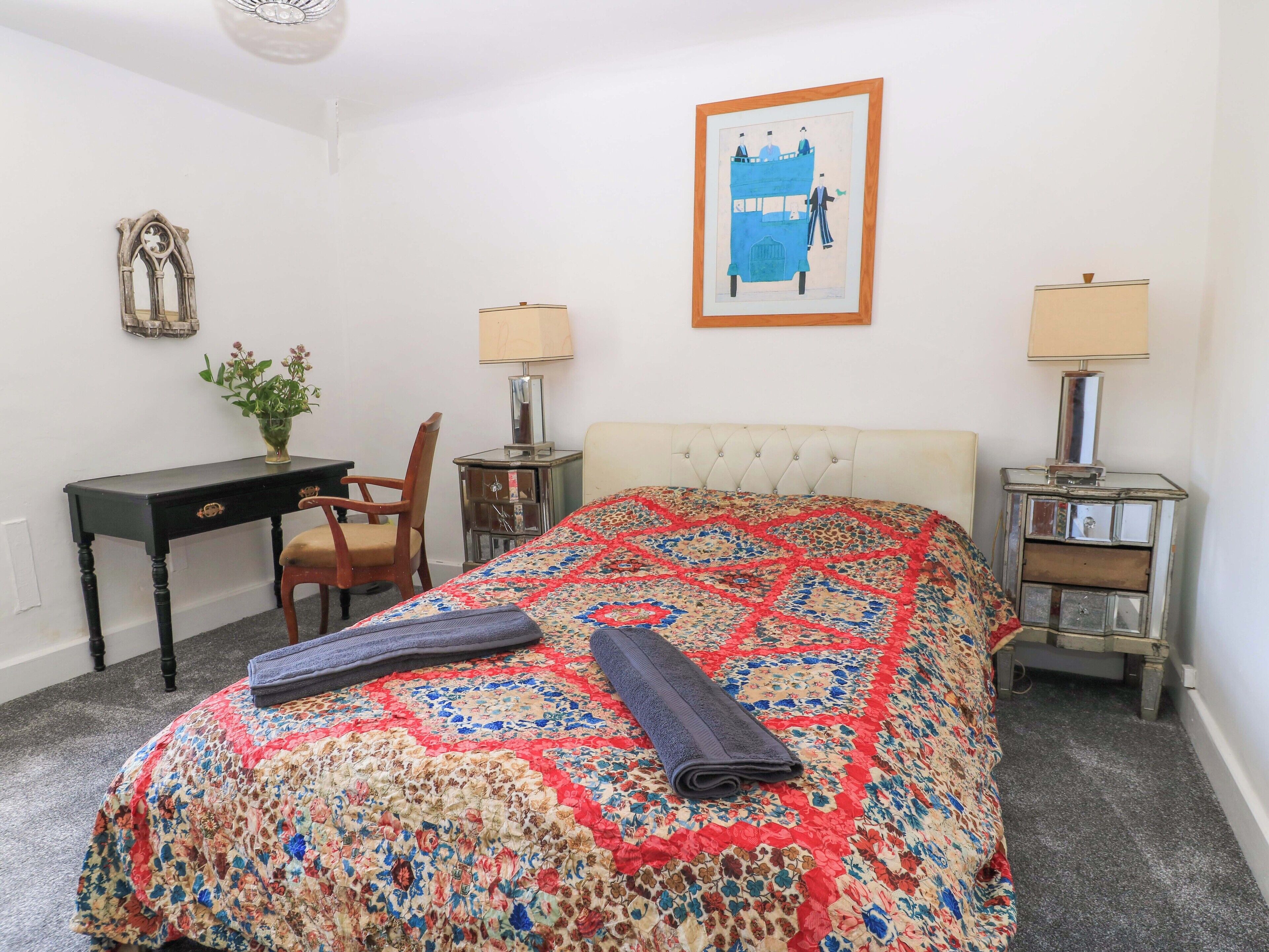 A hotel room with a bed, a desk, a chair, two bedside tables with lamps, a framed picture on the wall, and a decorative mirror.
