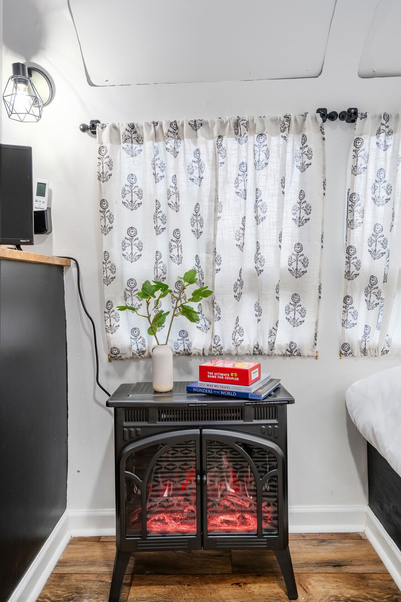 Une pièce de taille modeste comprenant un lit, une petite table avec des livres, une plante décorative et un poêle à bois.