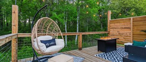 A wooden deck with a hanging chair, a small table with a fire feature, and patterned rugs.
