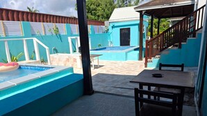 Un espace piscine avec une table et des chaises en bois, un coin salon couvert et un escalier menant à un bâtiment.