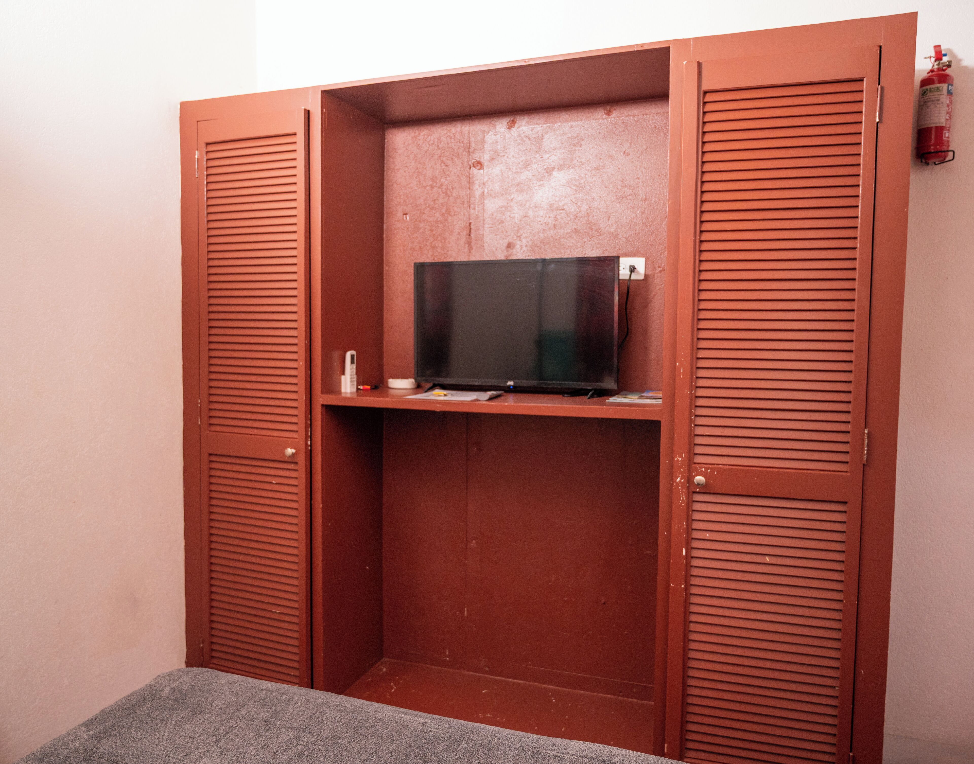 Habitación compacta con televisión integrada en un estante, un escritorio pequeño y un extintor de incendios fijado en la pared.