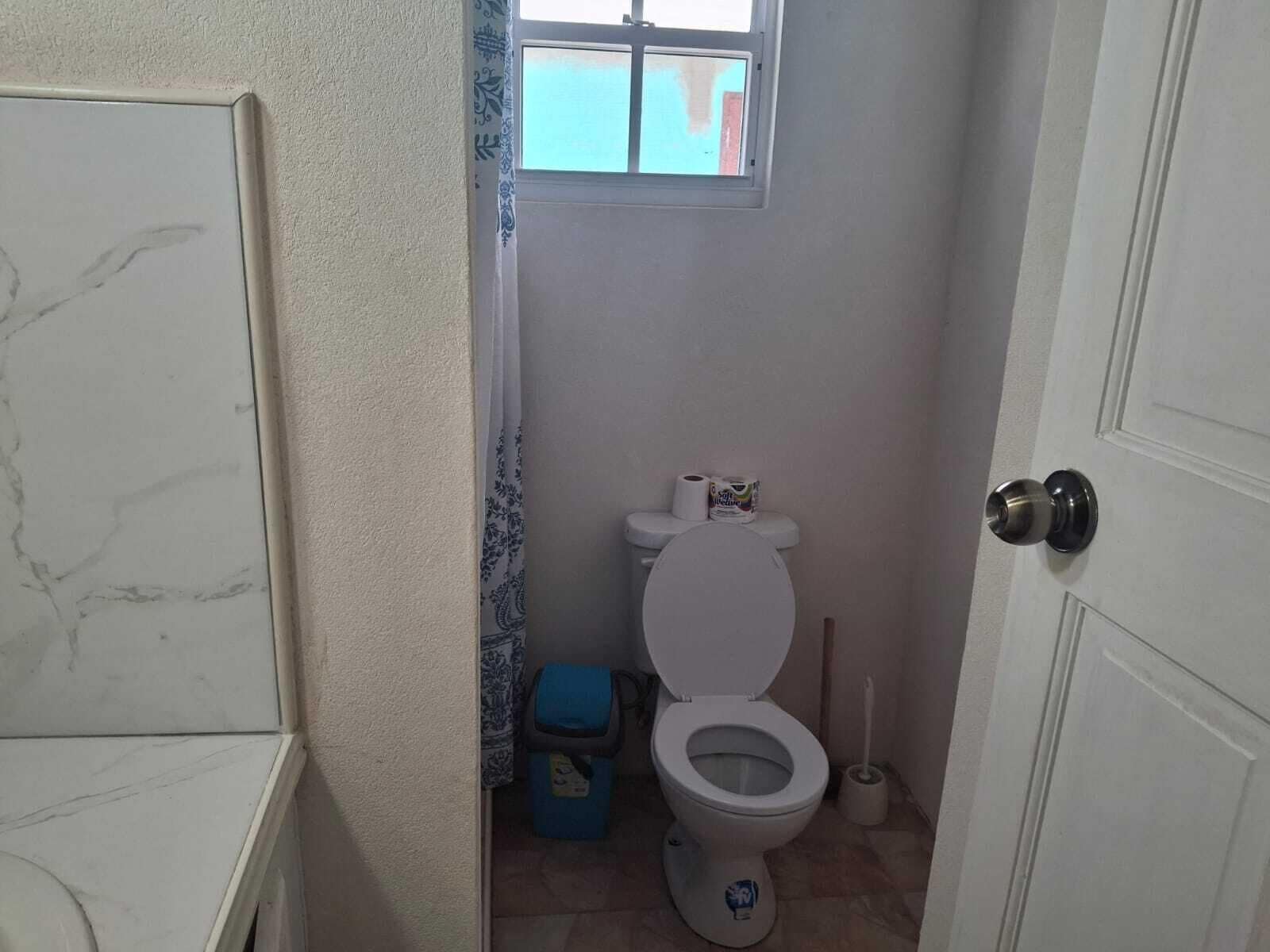 Une petite salle de bain avec des toilettes, une poubelle bleue et une fenêtre avec un rideau.