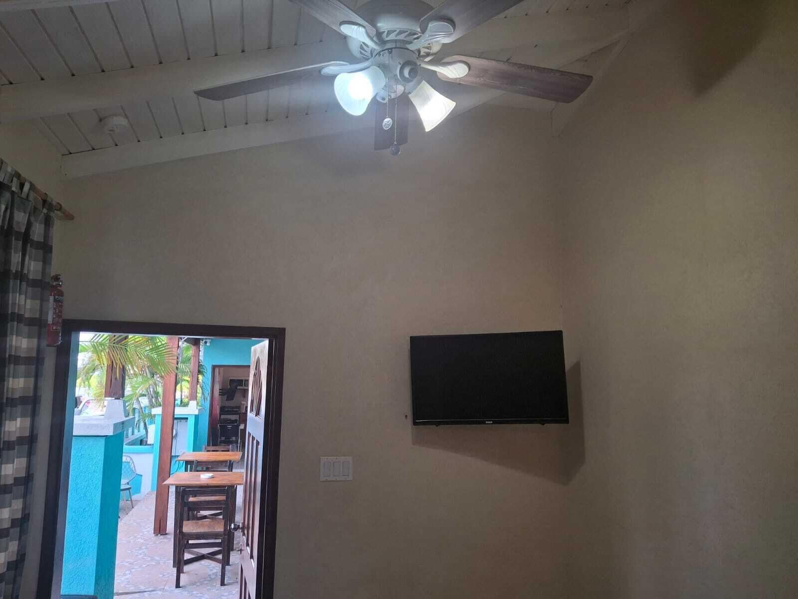 Ein Zimmer mit Deckenventilator, einem an der Wand befestigten Flachbildfernseher und einem Durchgang zu einem Außenbereich mit Tisch und Stühlen.