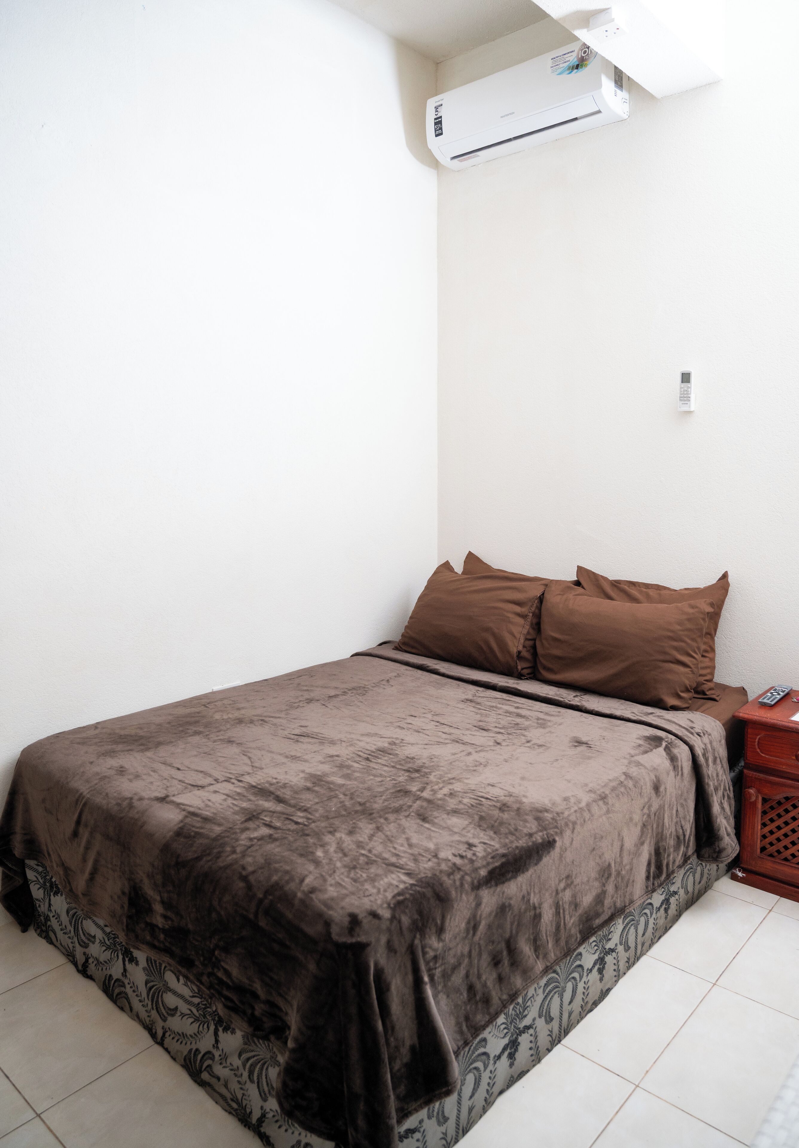 A single bed with a brown bedspread and two pillows, a small bedside table with a phone, and an air conditioning unit mounted on the wall.