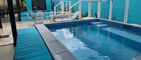 A swimming pool with a blue tiled edge, surrounded by a covered patio area with seating and a small table.