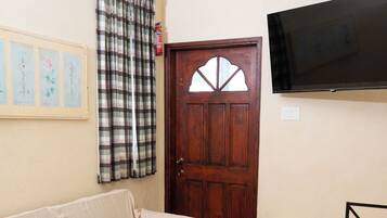 A room with a beige sofa, a wooden door, a television mounted on the wall, and a dining table with chairs.