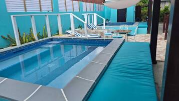 Una piscina con borda piastrellata blu, un solarium bianco e un'area salotto coperta con cuscini azzurri.