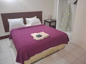 A neatly made bed with a purple bedspread and two white pillows. There is a small bedside table with a lamp and a floral curtain partially visible.
