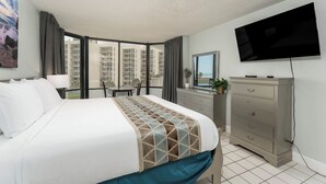 A hotel room with a large bed, a dresser, a TV, and a balcony with a view of high-rise buildings. - Spacious Beachfront Condo with Beautiful Views & Resort Amenities – Great Value! (South Padre Island)
