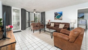 A spacious living room with a large sofa, armchairs, a coffee table, and a TV. There is a large window with curtains, a painting of a coastal scene, and a ceiling fan. - Spacious Beachfront Condo with Beautiful Views & Resort Amenities – Great Value! (South Padre Island)