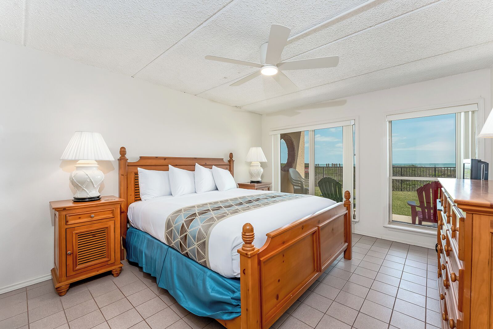 A bedroom with a wooden bed, two bedside tables with lamps, a ceiling fan, a large window with a view, and a sliding glass door leading to an outdoor area.