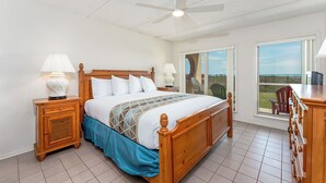 A bedroom with a wooden bed, two bedside tables with lamps, a ceiling fan, a large window with a view, and a sliding glass door leading to an outdoor area. - Suntide II 102 - Spacious Ground Floor Oceanfront Condo, Private Balcony, Pool, Spa, BBQ, Tennis and More (South Padre Island)