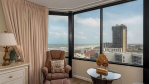 Una habitación con un ventanal grande que da a una playa y al paisaje urbano, un sillón mullido, una pequeña mesa redonda y un aparador blanco con una lámpara.