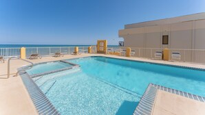A pool area with clear blue water, a white building with a balcony, and a view of the ocean.