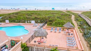 A pool area with lounge chairs and a thatched umbrella, overlooking a beach with umbrellas and a lifeguard stand.