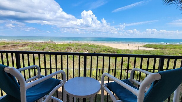 A balcony with two chairs and a small table, overlooking a beach with a wooden boardwalk and grassy areas.
