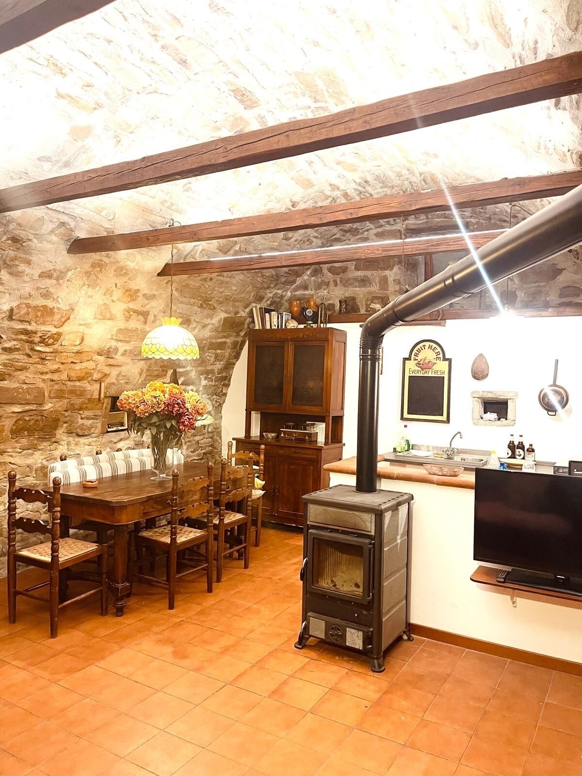 A rustic kitchen with a wood-burning stove, stone walls, and a dining area with wooden furniture.