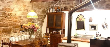 A rustic kitchen with a wood-burning stove, stone walls, and a dining area with wooden furniture.