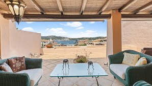 Un espacio de estar al aire libre cubierto con muebles de mimbre y una mesa de vidrio, con vistas a un paisaje pintoresco de un pueblo costero.