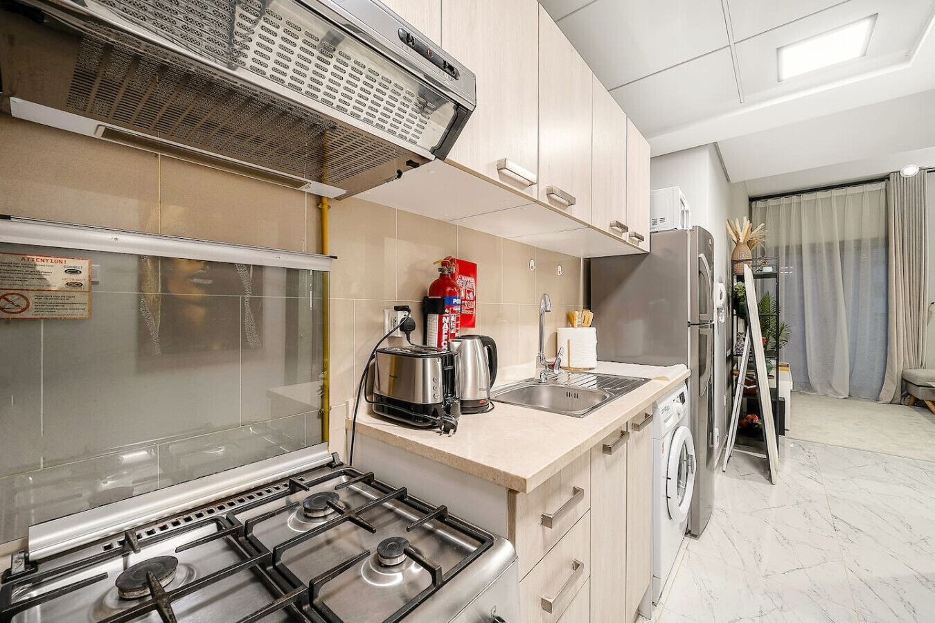 Cocina moderna con estufa a gas, tostadora, hervidor y horno empotrado. Se observa un lavamanos, un refrigerador y una ventana con vistas.