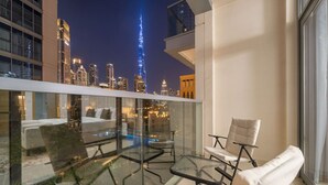 A balcony with a glass barrier, a white sofa, a glass table, and a chair, overlooking a cityscape with illuminated buildings and a prominent tower. - FIRST CLASS | 1BR | Burj Khalifa & Pool View (Downtown)
