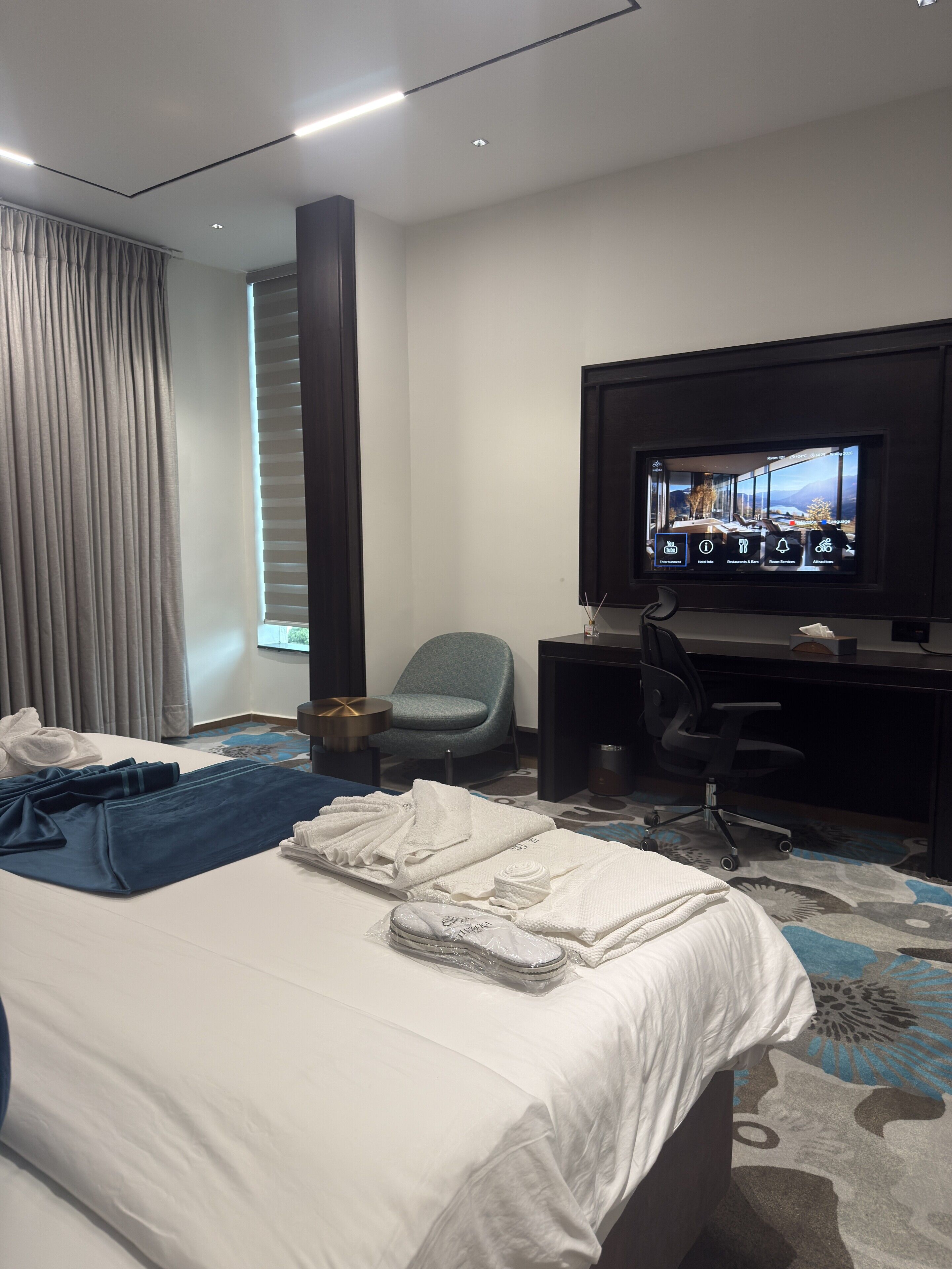 A hotel room with a bed, a desk with a computer, a chair, and a television displaying room service options.
