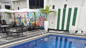 Une piscine dont le bord est recouvert de carreaux bleus, entourée d’une clôture blanche, et dont un mur est décoré d’une fresque.