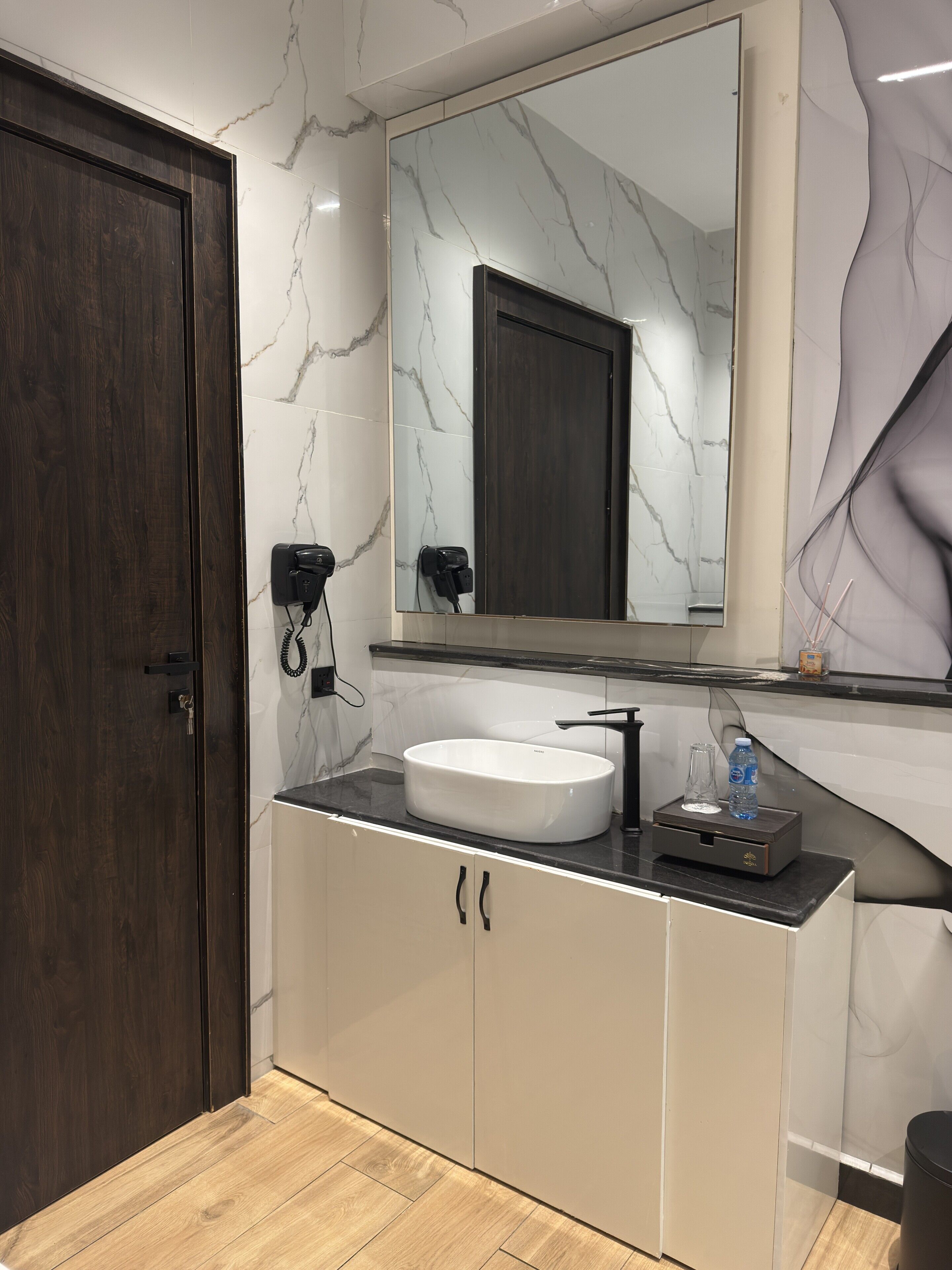 A modern bathroom with a marble wall, a wooden door, a white sink, and a large mirror.