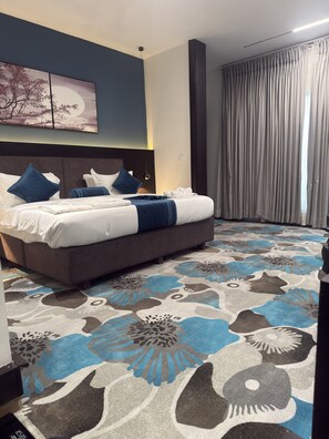 A hotel room with a large bed, a headboard, a wall-mounted artwork, a window with curtains, and a patterned carpet. - THE THRIBEKA INTERNATIONAL HOTEL (Asaba)