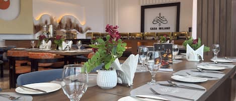 A long dining table set with white tableware and a vase of flowers, in a room with a high ceiling and modern decor.