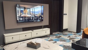 A modern hotel room with a large flat-screen TV mounted on the wall, a marble dining table, two blue chairs, a white console table, and a patterned carpet. - THE THRIBEKA INTERNATIONAL HOTEL (Asaba)
