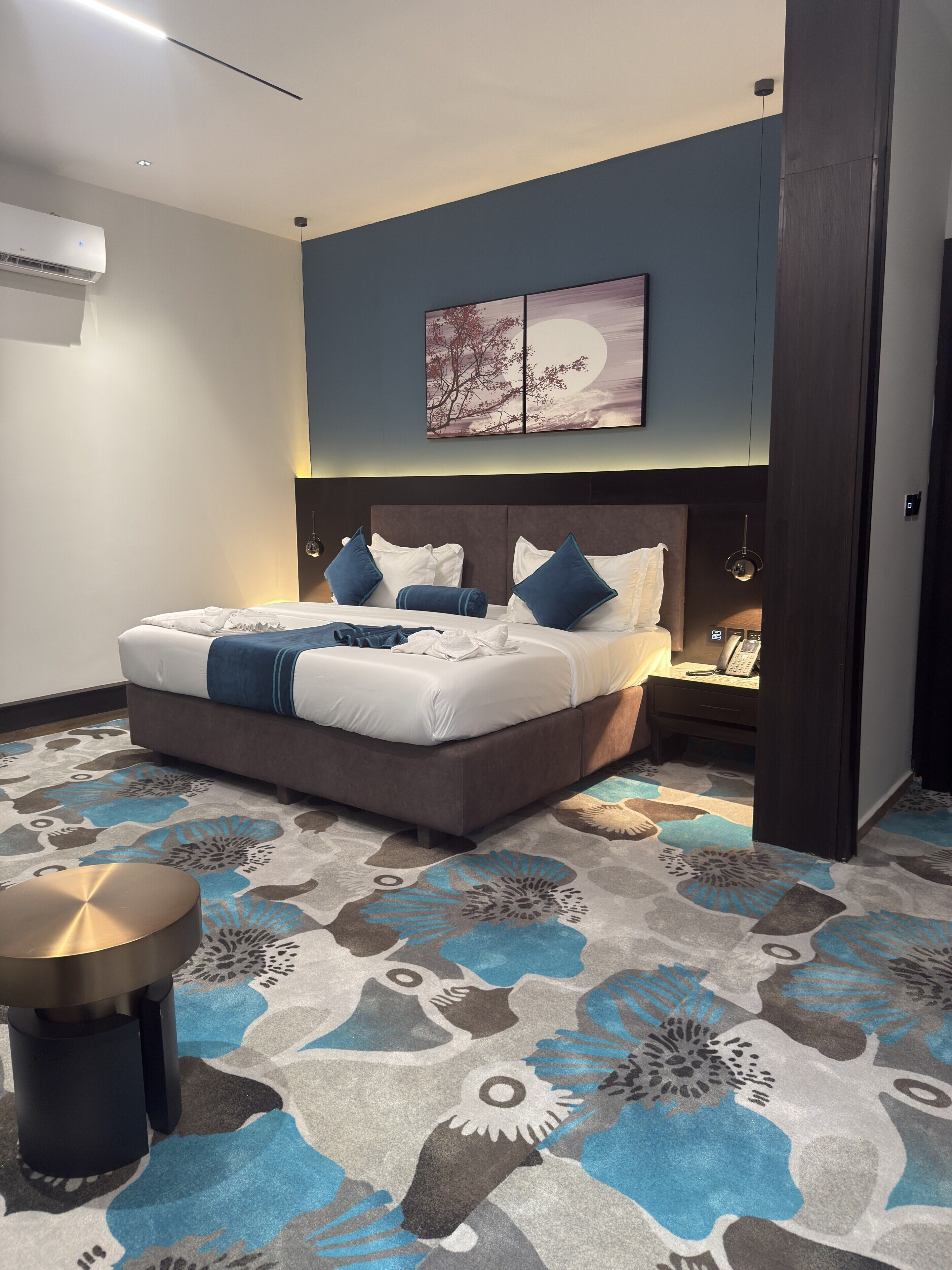 Habitación de hotel moderna con una cama grande, una mesita de noche con lámpara, un teléfono en la pared y un cuadro decorativo sobre la cama.