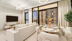 A modern living room with a large flat-screen TV, a white sofa, a dining table set for four, and a large window with a city view. - Stylish 2BR — Creek Harbour Views Near Yacht Club & Dining Spots (Dubai Creek Harbour)