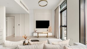 A modern hotel room with a large bed, a flat-screen TV mounted on the wall, a small table with a vase of flowers, and a view of the city through the window. - Stylish 2BR — Creek Harbour Views Near Yacht Club & Dining Spots (Dubai Creek Harbour)