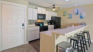 A modern kitchen with white cabinets, a black refrigerator, a microwave, and a stove. There are three bar stools and a trash can. - Cozy Pet-Friendly Condo w/ Pool (Osage Beach)