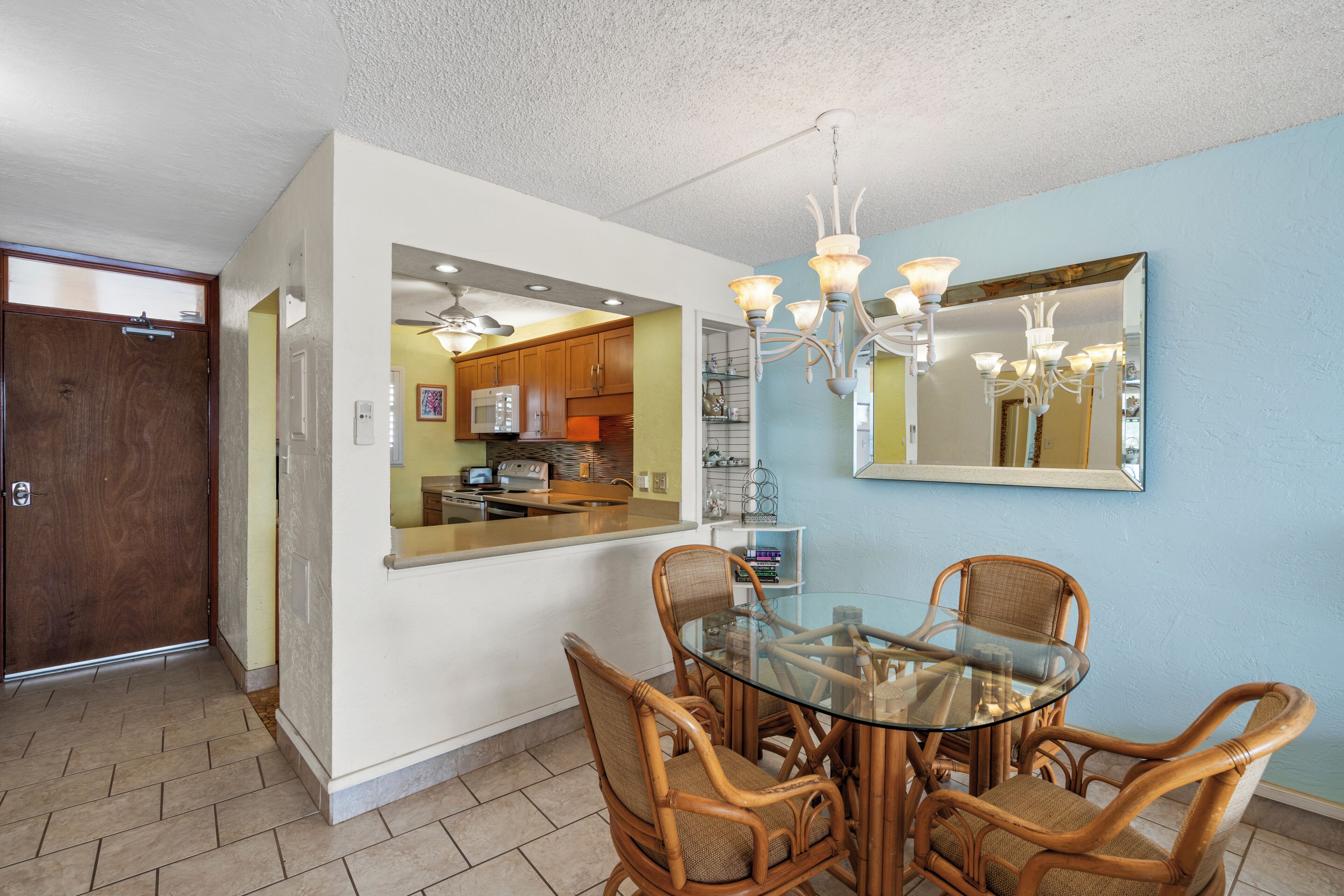 A dining area with a glass table and wicker chairs, a kitchen visible through an open door, and a chandelier above.