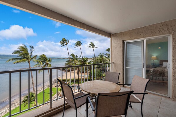 A balcony with a view of the ocean, palm trees, and a table with chairs.