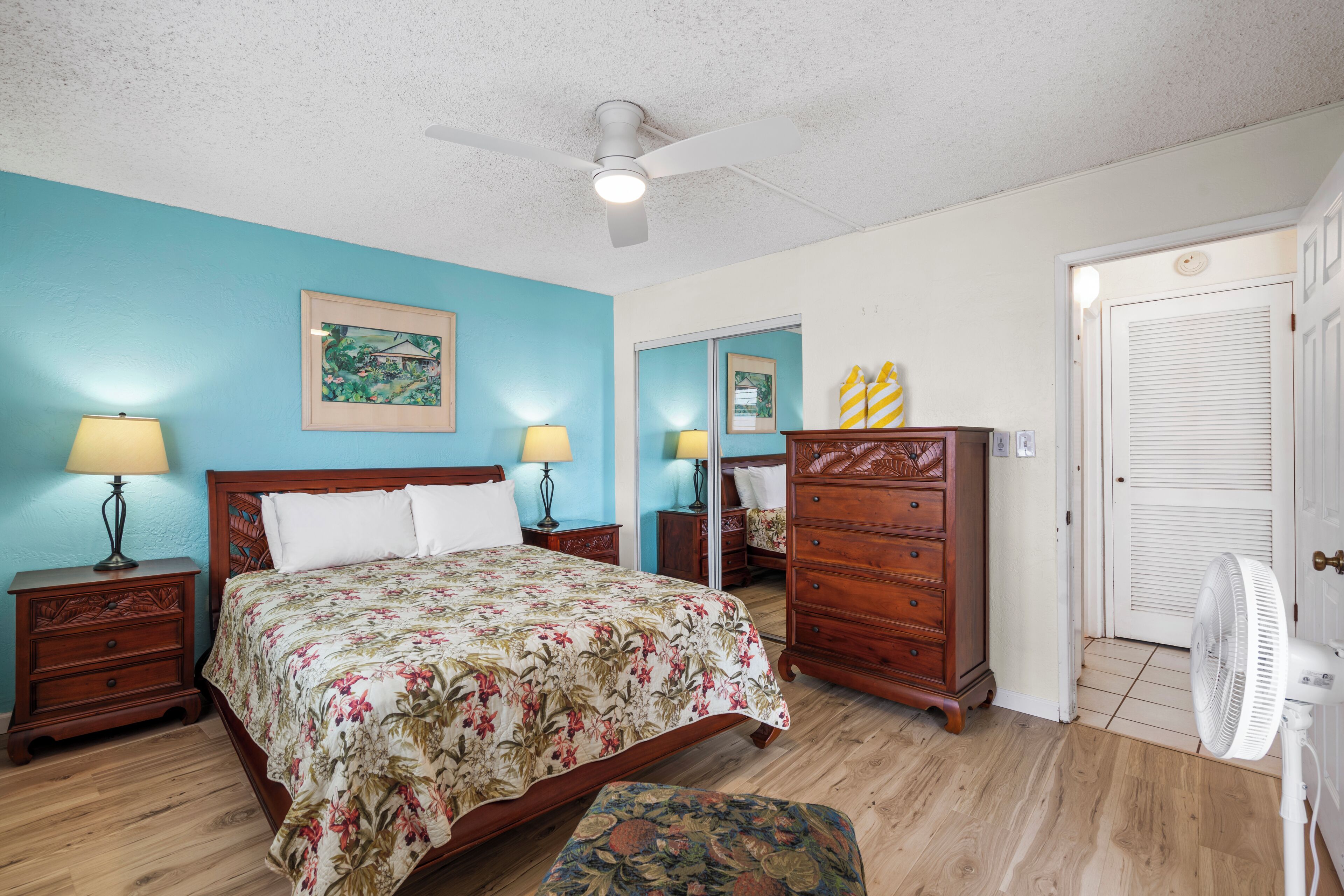A bedroom with a bed, nightstands, a dresser, a fan, and a door.