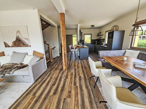 A modern interior with a wooden floor, a dining area with a wooden table, a kitchen, and a living area with a sofa and a fur throw.