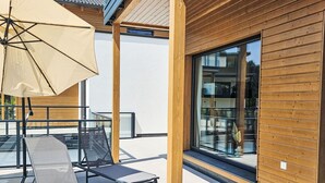 A modern outdoor seating area with lounge chairs and a large umbrella, wooden paneling on the building, and a large glass window.