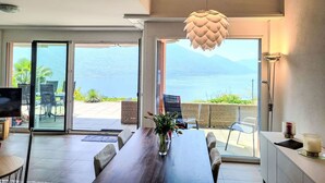 A modern dining area with a wooden table, chairs, and a vase of flowers. Large sliding glass doors lead to an outdoor patio with a view of a lake and mountains. - Nice apartment for 4 guests with WIFI, TV, patio, pets allowed and panoramic view (Tenero)