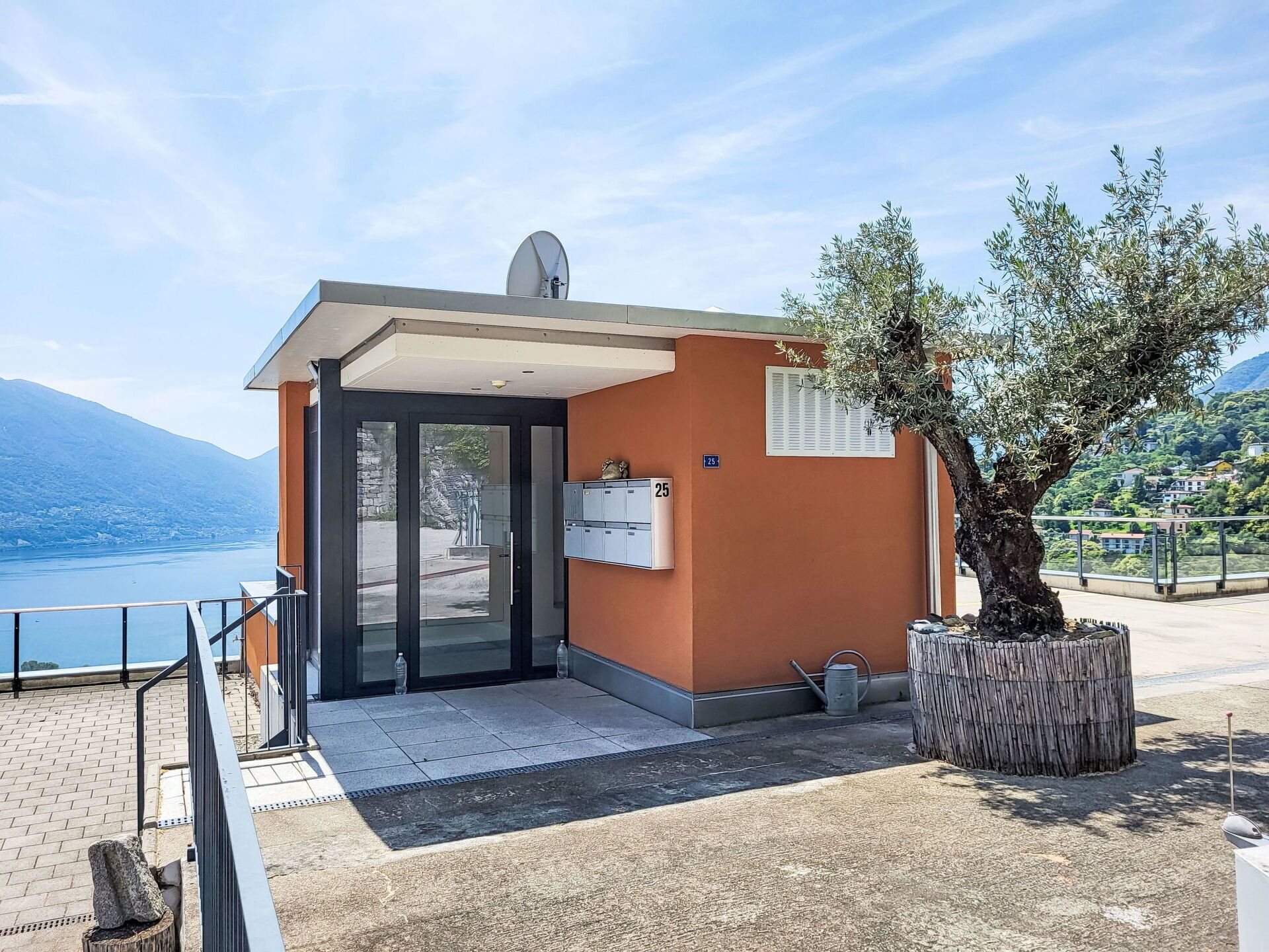Un petit bâtiment avec une porte d’entrée en verre, une antenne parabolique et une vue sur un lac et des montagnes.